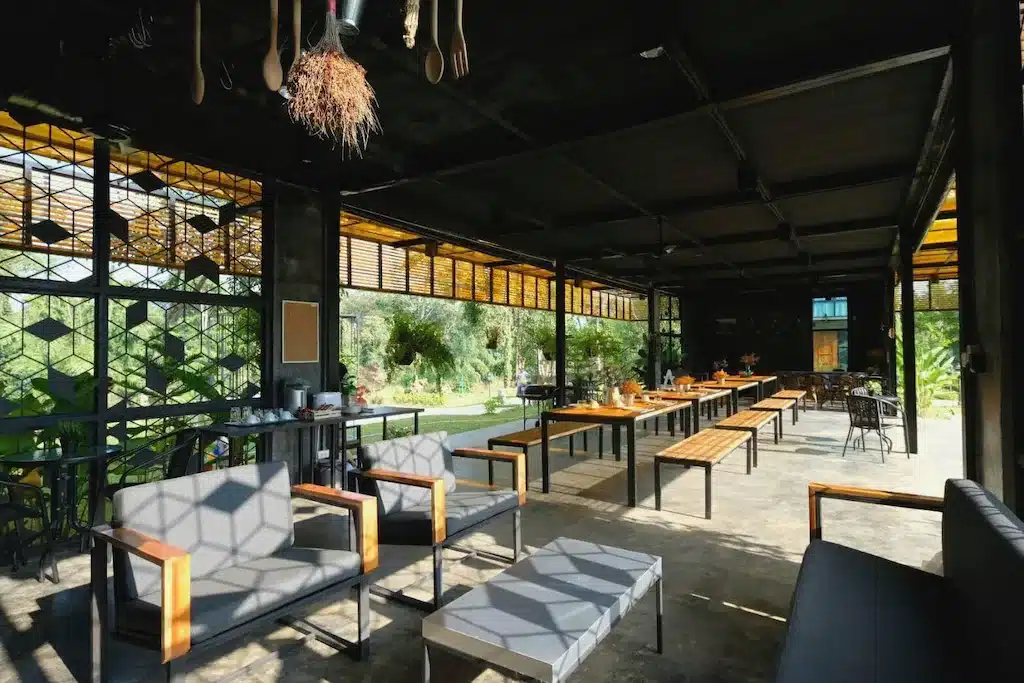 คาเฟ่กลางแจ้งพร้อมเฟอร์นิเจอร์ ที่พักกาญจนบุรี โลหะและไม้สมัยใหม่ หน้าต่างบานใหญ่ และแสงธรรมชาติที่ทอดเงาเรขาคณิตลงบนพื้น; มีต้นไม้สีเขียวมองเห็นได้จากภายนอก—เหมาะสำหรับแขกจากที่พักกาญจนบุรีหรือรีสอร์ทกาญจนบุรีใกล้เคียง.