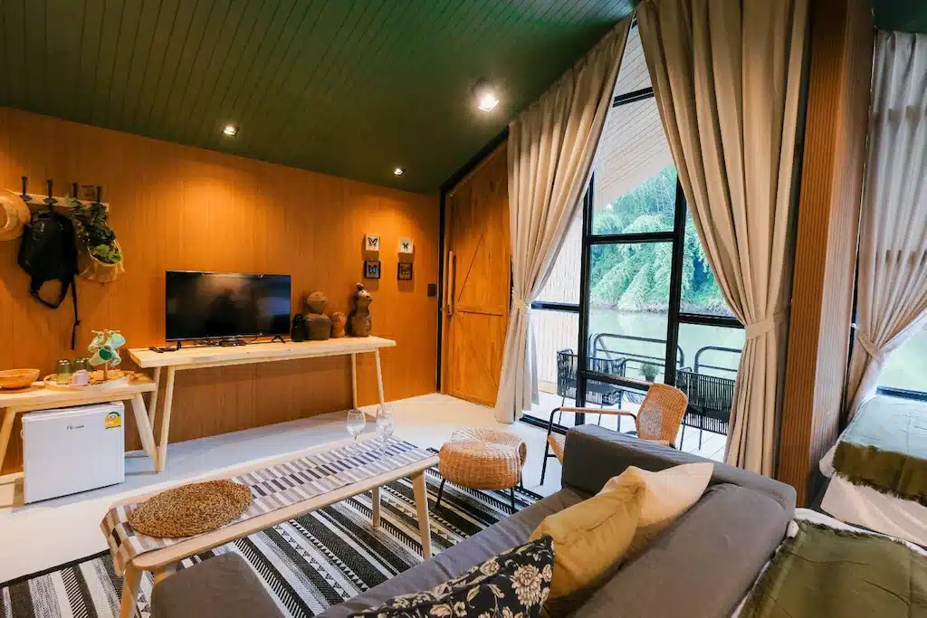 ห้องนั่งเล่น ที่พักกาญจนบุรี สไตล์โมเดิร์นพร้อมผนังไม้, โซฟา, ทีวี, ตู้เย็นขนาดเล็ก, และหน้าต่างบานใหญ่พร้อมผ้าม่านที่มองเห็นระเบียงและต้นไม้เขียวขจี—เหมาะสำหรับการผ่อนคลายที่ ที่พักกาญจนบุรีริมน้ำ.