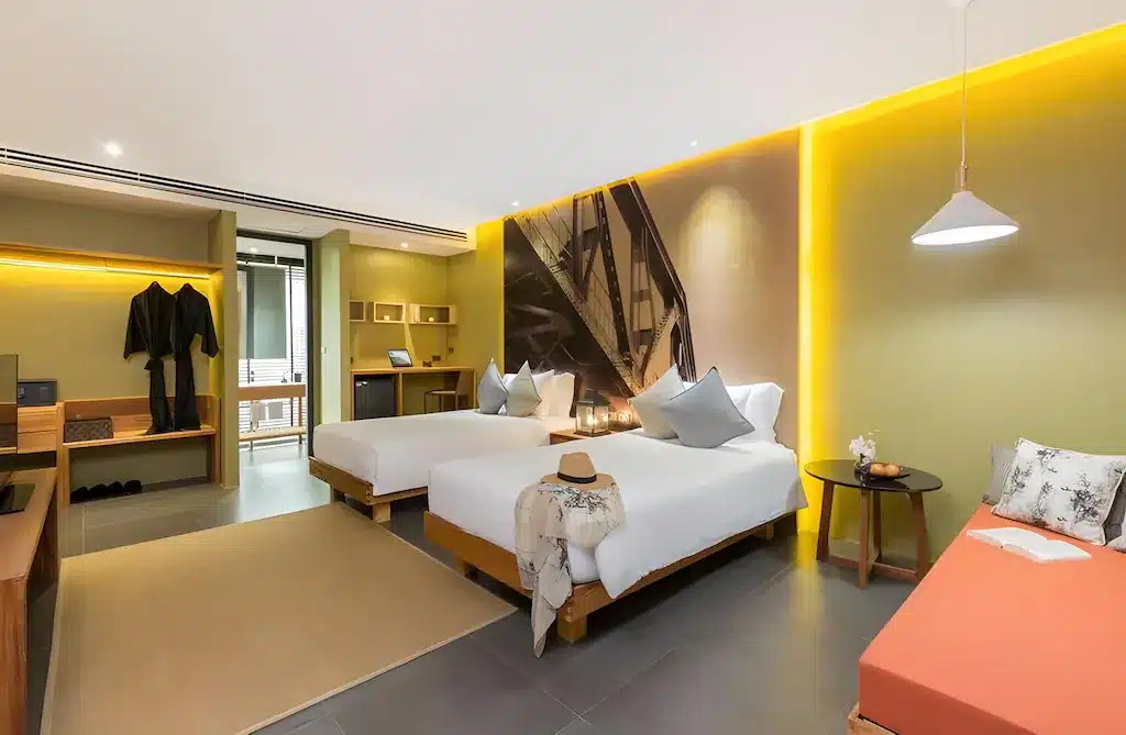 ห้องพักโรงแรมสมัยใหม่ที่รีสอร์ท ที่พักกาญจนบุรี กาญจนบุรี พร้อมเตียงคู่สองเตียง, โซฟา, ตู้เสื้อผ้าแบบเปิด, ภาพศิลปะบนผนัง, พื้นที่ทำงาน, และแสงสว่างอบอุ่นเพื่อการพักผ่อนอย่างผ่อนคลาย.