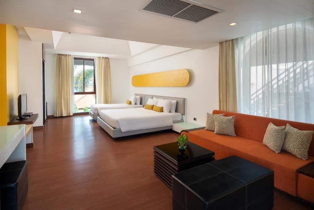 ห้องพักในโรงแรมทันสมัยพร้อมเตียงคู่ 2 เตียง โซฟาสีส้ม โต๊ะกาแฟ ทีวี และหน้าต่างบานใหญ่พร้อมผ้าม่านที่เปิดรับแสงธรรมชาติ เหมาะสำหรับผู้ที่กำลังมองหาที่พักพัทยาราคาถูกและการพักผ่อนอย่างสะดวกสบายในที่พักของพัทยา.