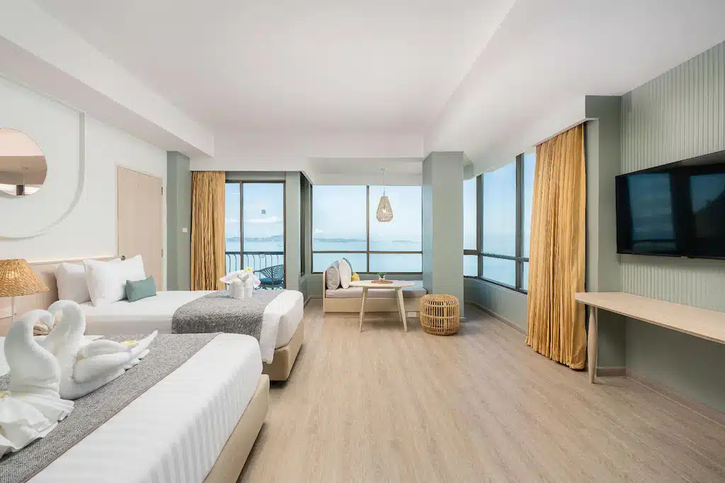 ห้องพักในโรงแรมทันสมัยพร้อมเตียง 2 เตียง ทีวีติดผนัง พื้นที่นั่งเล่นริมหน้าต่างบานใหญ่ พื้นไม้สีอ่อน และวิวทะเลที่สวยงาม เหมาะสำหรับผู้ที่กำลังมองหาพัทยาราคาถูกสำหรับการพักผ่อนอย่างผ่อนคลาย.