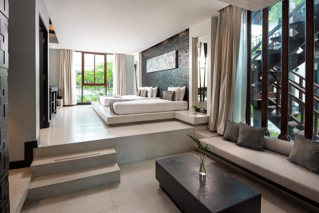 ห้องพักในโรงแรมทันสมัยพร้อมเตียงยกสูง หน้าต่างบานใหญ่ ผนังเบา และม้านั่งในตัวพร้อมเบาะรองนั่ง แสงธรรมชาติเข้ามาเติมเต็มพื้นที่ เหมาะสำหรับผู้ที่มองหาที่พักพัทยาราคาถูกอย่างมีสไตล์และสะดวกสบาย.