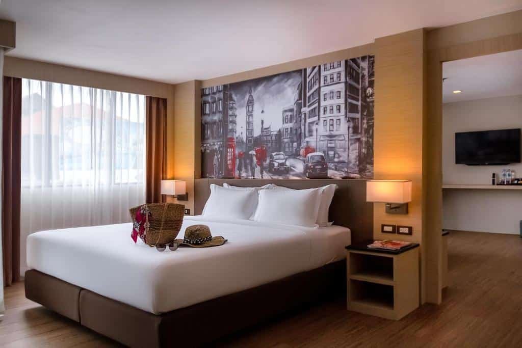 ห้องพักโรงแรมทันสมัยในที่พักของพัทยาพร้อมเตียงขนาดใหญ่ โต๊ะข้างเตียง โคมไฟ ภาพจิตรกรรมฝาผนัง หน้าต่างพร้อมผ้าม่าน รวมถึงบริเวณโต๊ะทำงานพร้อมทีวีและเครื่องดื่ม - เหมาะสำหรับผู้ที่กำลังมองหาพัทยาราคาถูก.