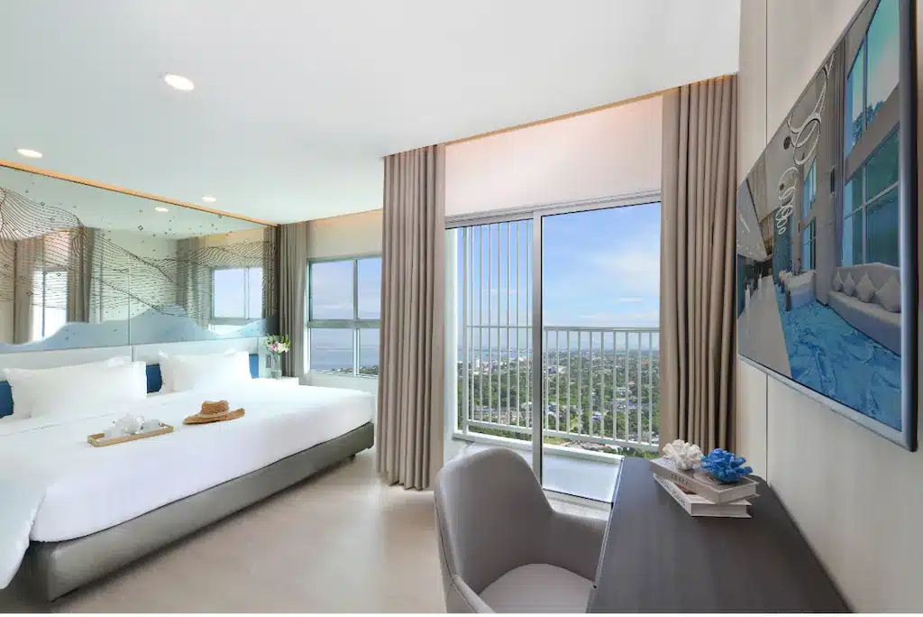 ห้องพักในโรงแรมทันสมัยพร้อมเตียงขนาดใหญ่ โต๊ะ เก้าอี้ และทีวีติดผนัง ประตูหน้าต่างและกระจกที่มองเห็นวิวท้องฟ้าสีครามและทิวทัศน์ เหมาะสำหรับผู้ที่กำลังมองหาที่พักพัทยาราคาถูกในบรรยากาศสบายๆ.