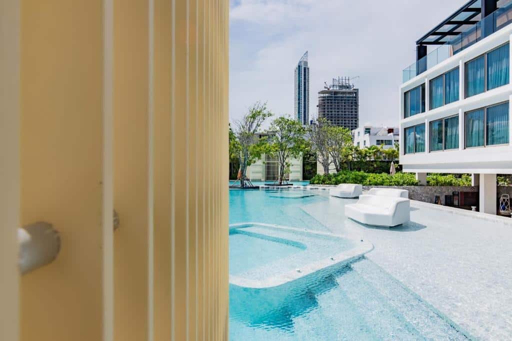 สระว่ายน้ำกลางแจ้งพร้อมเก้าอี้เลานจ์สีขาว อาคารทันสมัยทางด้านขวา เส้นขอบฟ้าของเมืองและการก่อสร้างที่อยู่เบื้องหลัง เป็นจุดที่น่าสนใจสำหรับ ที่พักพัทยาราคาถูก มีกรอบบางส่วนด้วยแท่งสีเหลืองแนวตั้งทางด้านซ้าย.