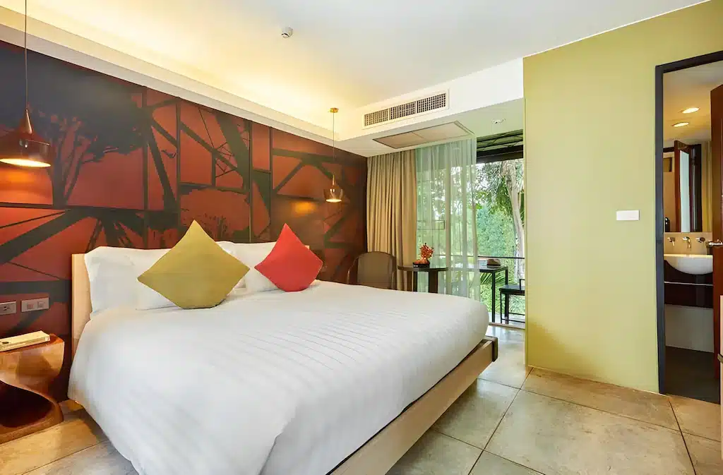 ห้องพักโรงแรมสมัยใหม่พร้อมเตียงคู่ หมอนสามใบ ภาพจิตรกรรมฝาผนัง  ที่พักกาญจนบุรี ไฟแขวน โต๊ะและเก้าอี้ ระเบียงที่สามารถเข้าถึงได้ และห้องน้ำที่มองเห็นได้ผ่านประตูที่เปิดอยู่—เหมาะสำหรับการเข้าพักของคุณที่ ที่พักกาญจนบุรีริมน้ำ.
