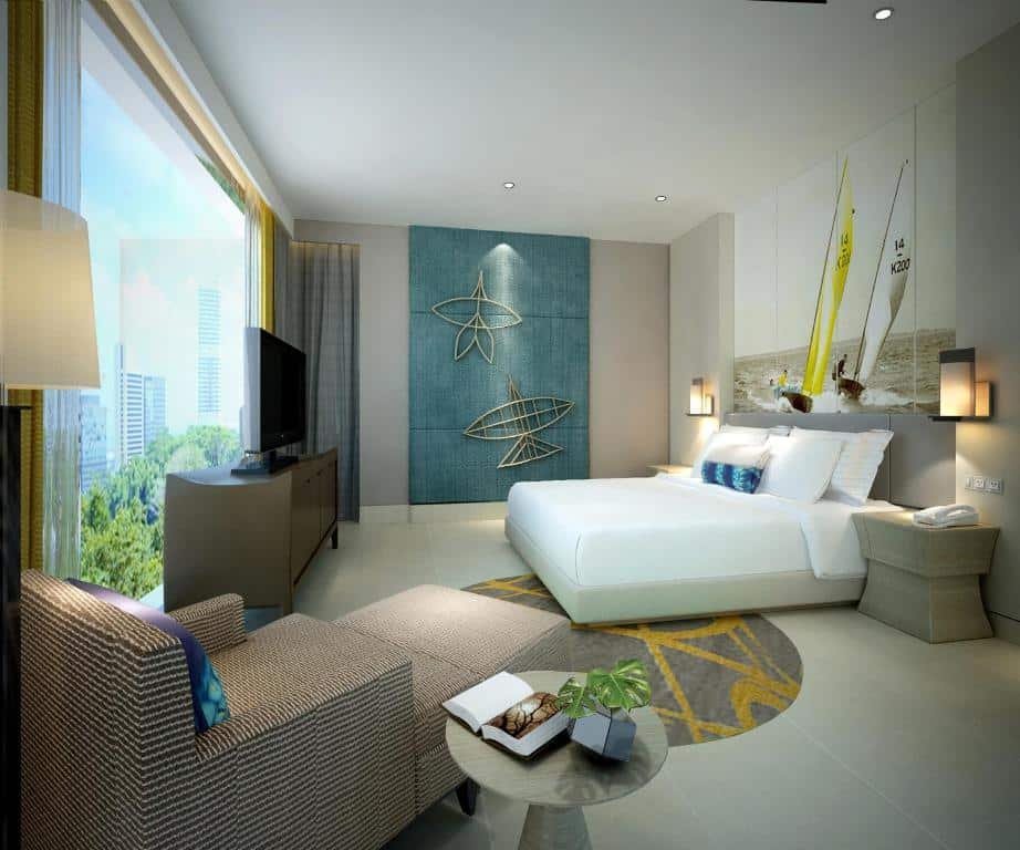 ห้องพักในโรงแรมทันสมัยพร้อมเตียงขนาดใหญ่ เก้าอี้ โทรทัศน์ และวิวเมืองผ่านหน้าต่างบานสูงจากพื้นจรดเพดาน งานศิลปะบนผนังธีมทะเลและการตกแต่งแบบเรียบง่ายสร้างบรรยากาศผ่อนคลาย เหมาะสำหรับผู้ที่มองหาที่พักในพัทยาที่มีสไตล์และสะดวกสบาย.