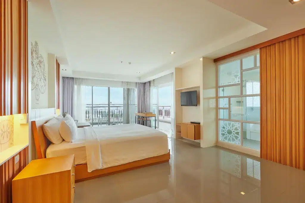 ห้องพักในโรงแรมกว้างขวางพร้อมเตียงขนาดใหญ่ เฟอร์นิเจอร์ไม้ ทีวีจอแบน ห้องน้ำผนังกระจก และระเบียงพร้อมวิวทะเลผ่านประตูบานเลื่อนกระจก เป็นตัวเลือกในอุดมคติสำหรับการพักในพัทยาพร้อมความสะดวกสบายที่น่าทึ่ง.