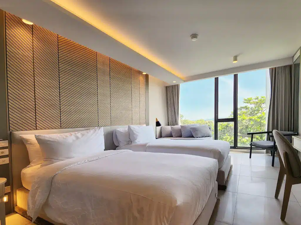ห้องพักในโรงแรมทันสมัยพร้อมเตียงขนาดใหญ่ 2 เตียง การตกแต่งที่เป็นกลาง โต๊ะและเก้าอี้ และหน้าต่างสูงจากพื้นจรดเพดานที่มองเห็นความเขียวขจี เหมาะสำหรับผู้ที่กำลังมองหาพัทยาราคาถูกในบรรยากาศที่เงียบสงบ.