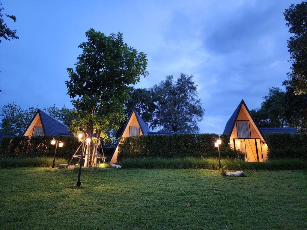 กระท่อม A-frame ขนาดเล็กสามหลังที่มีหน้าต่างสว่างตั้งอยู่หลังพุ่มไม้บนสนามหญ้าในยามพลบค่ำ โดยมีต้นไม้และเสาไฟส่องสว่างอยู่ด้านหน้า—เป็นที่พักที่อบอุ่นเหมาะสำหรับผู้ที่มองหา ที่พักกาญจนบุรี หรือรีสอร์ทกาญจนบุรี.