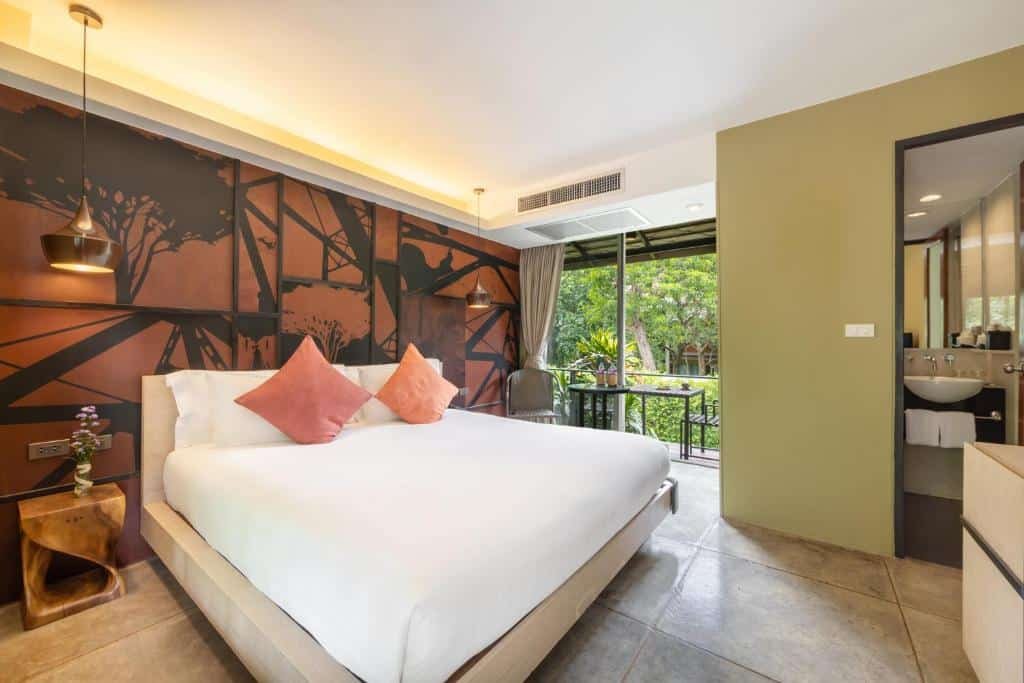 ห้องพักโรงแรมสมัยใหม่พร้อมเตียงขนาดใหญ่ หมอนสีชมพูสองใบ ผนังตกแต่งด้วยภาพวาดฝาผนัง ระเบียงเล็กพร้อมวิวสวน และห้องน้ำแบบเปิดโล่ง—เหมาะสำหรับผู้ที่ต้องการประสบการณ์การพักผ่อนที่ผ่อนคลาย ที่พักกาญจนบุรี หรือ รีสอร์ทกาญจนบุรี.