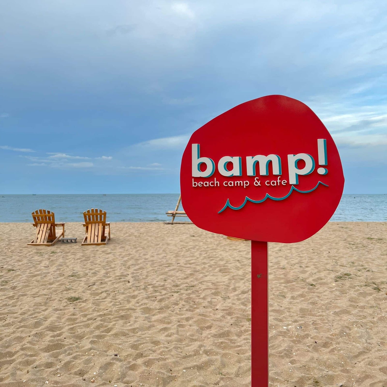 ป้ายสีแดงเขียนว่า "bamp! beach camp & cafe" บนหาดทรายพร้อม ท่องเที่ยวหัวหิน เก้าอี้ไม้เปล่าสองตัวหันหน้าไปทางทะเลภายใต้ท้องฟ้าที่มีเมฆมาก สถานที่เงียบสงบที่เที่ยวธรรมชาติหัวหินเหมาะสำหรับผู้ที่แสวงหาความสงบที่ท่องเที่ยวหัวหิน.