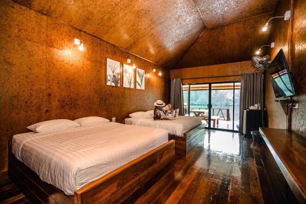 ห้องไม้สักสองเตียงคู่ที่ ที่พักกาญจนบุรีริมน้ำ, มีผู้หนึ่งกำลังผ่อนคลายอยู่บนเตียง; หน้าต่างบานใหญ่ล้อมรอบด้วยต้นไม้เขียวขจี, พร้อมด้วยโทรทัศน์, โต๊ะทำงาน, และศิลปะติดผนังเพื่อการพักผ่อนที่แสนสบาย.
