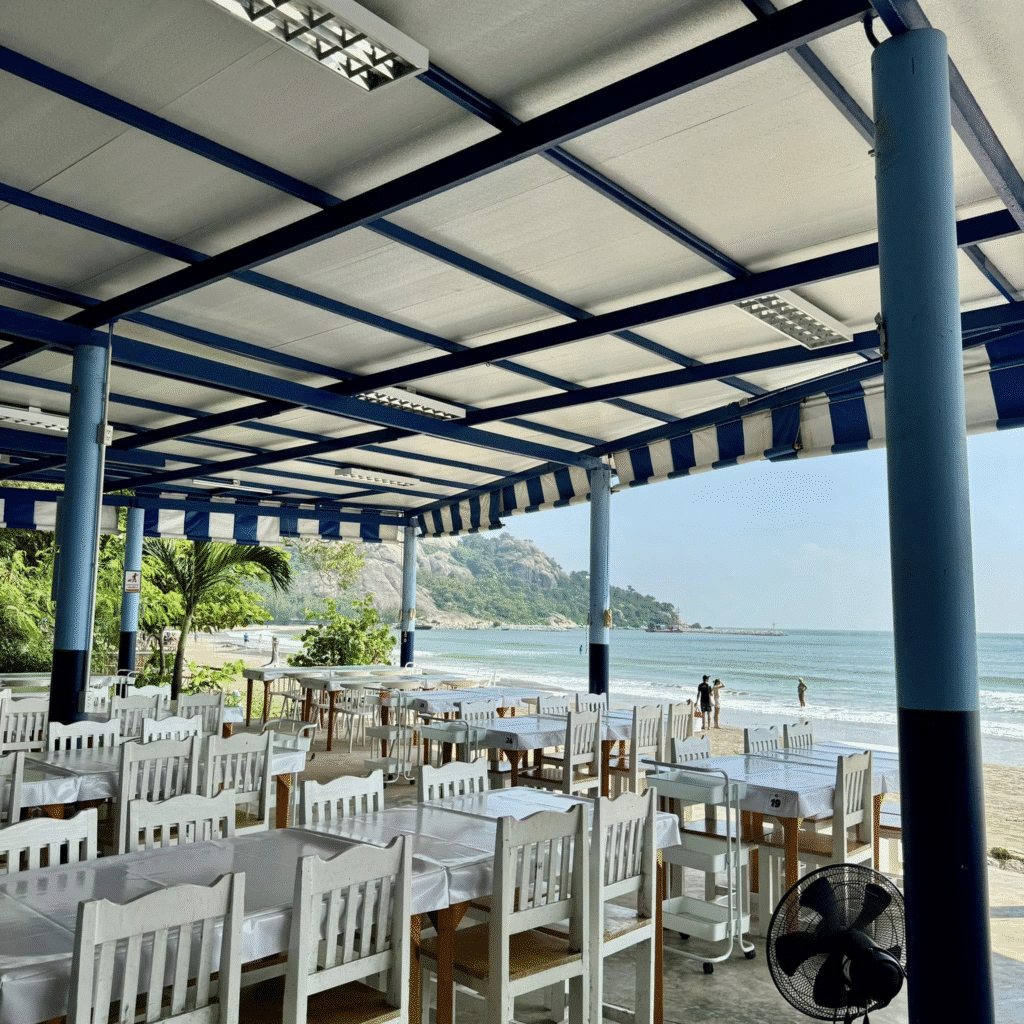 ร้านอาหารแบบเปิดโล่งริมชายหาดพร้อมโต๊ะและเก้าอี้สีขาวใต้ร่มไม้สีฟ้าขาว มองเห็นทะเลและแนวหาดทราย เป็นจุดพักผ่อนเพื่อเพลิดเพลินกับที่ ที่ท่องเทียวหัวหิน เที่ยวธรรมชาติหัวหินและเสน่ห์ของสถานที่ท่องเที่ยวหัวหิน.