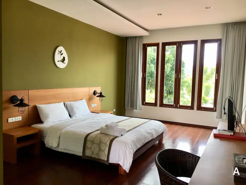 ห้องนอนสมัยใหม่ที่รีสอร์ทกาญจนบุรี พร้อมเตียงคู่, ผนังสีเขียว, หน้าต่างใหญ่สามบานพร้อมผ้าม่าน, พื้นไม้, และโต๊ะพร้อมเก้าอี้—ที่พักริมน้ำ ที่พักกาญจนบุรี กาญจนบุรีที่เหมาะสำหรับการพักผ่อนของคุณ.