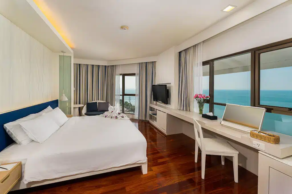 ห้องพักในโรงแรมทันสมัยพร้อมเตียงขนาดใหญ่ โต๊ะทำงาน ทีวี พื้นไม้ และหน้าต่างที่มองเห็นมหาสมุทร ดอกไม้และผ้าเช็ดตัวลายหงส์เพิ่มเสน่ห์ เหมาะสำหรับผู้ที่แสวงหาที่พักในพัทยาทั้งในด้านความสะดวกสบายและมีสไตล์.