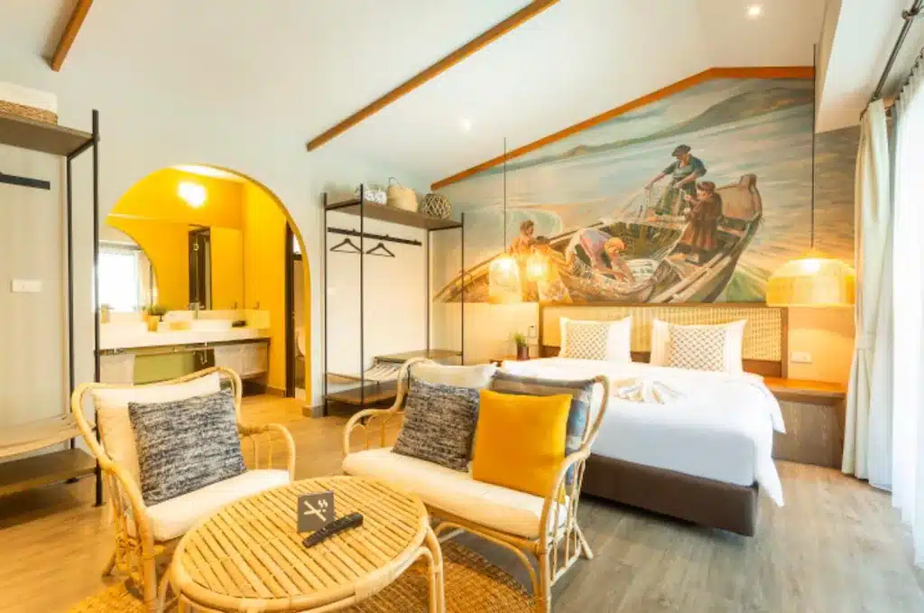 ห้องพักในโรงแรมที่สว่างสดใส ท่องเที่ยวสตูล ใจกลางท่องเที่ยวหัวหิน ประกอบไปด้วยเตียงคู่ เก้าอี้หวาย 2 ตัว โต๊ะกลม ตู้เสื้อผ้าแบบเปิด และภาพคนกำลังตกปลาบนฝาผนัง เหมาะสำหรับการพักผ่อนที่เที่ยวธรรมชาติหัวหินของคุณ.