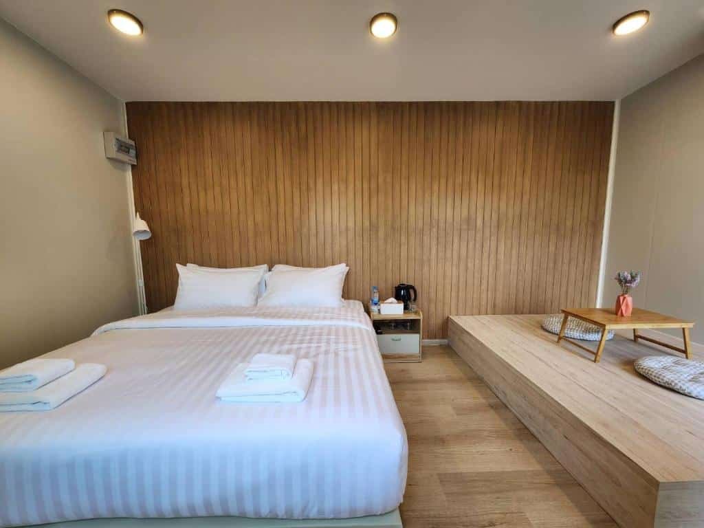 ห้องพักโรงแรมสมัยใหม่ที่ ที่พักกาญจนบุรีริมน้ำ ประกอบด้วยเตียงคู่, ผ้าเช็ดตัวพับไว้, ผนังไม้ตกแต่ง, โต๊ะข้างเตียงพร้อมกาต้มน้ำ, และมุมนั่งเล่นสบาย ๆ บนแพลตฟอร์มไม้พร้อมเบาะรองนั่งและโต๊ะเล็ก ๆ. ที่พักกาญจนบุรี 
