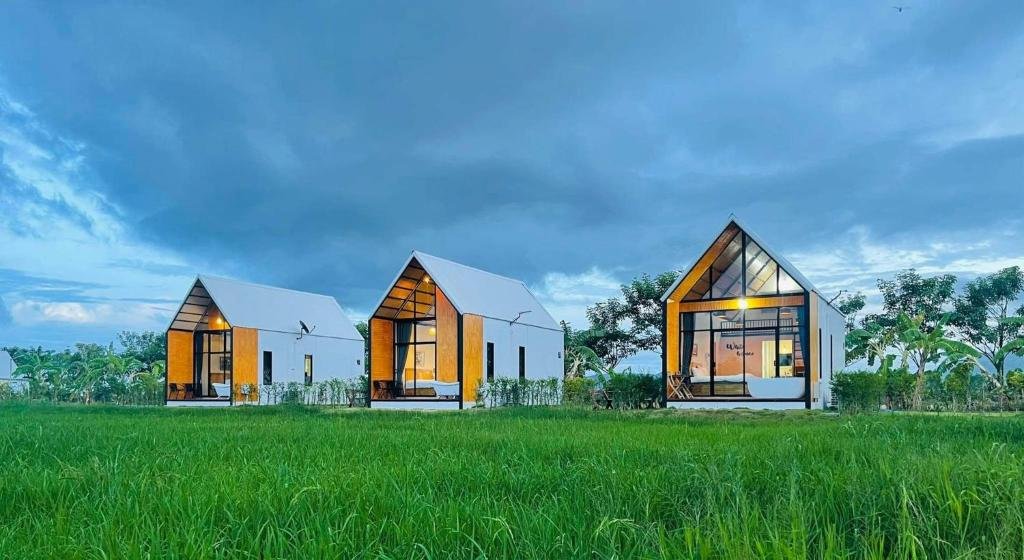 กระท่อมสไตล์โมเดิร์น ที่พักกาญจนบุรี และมินิมอลสามหลังที่มีหน้าต่างบานใหญ่ตั้งเรียงกันบนทุ่งหญ้าสีเขียวใต้ท้องฟ้าที่มีเมฆครึ้ม—ที่พักริมน้ำกาญจนบุรีที่เหมาะสำหรับผู้ที่ต้องการความสงบและสไตล์ท่ามกลางธรรมชาติ.