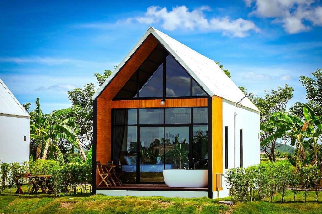 บ้านพักตากอากาศสไตล์โมเดิร์นข ที่พักกาญจนบุรี นาดเล็กที่มีหน้าต่างกระจกขนาดใหญ่ ผนังภายนอกสีขาว และหลังคาทรงจั่ว ตั้งอยู่ท่ามกลางธรรมชาติสีเขียวและที่นั่งกลางแจ้ง ท่ามกลางท้องฟ้าสีครามและเมฆขาว.