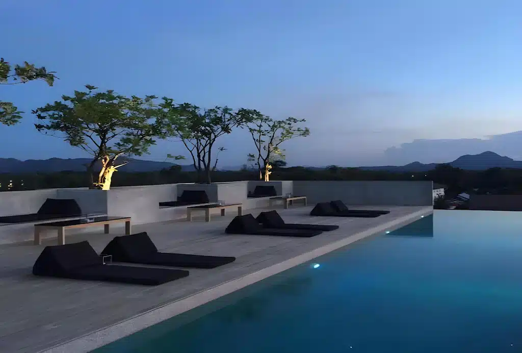 สระว่ายน้ำอินฟินิตี้ ที่พักกาญจนบุรี บนดาดฟ้าสมัยใหม่พร้อมเก้าอี้เลานจ์สีดำ ต้นไม้ในกระถาง และวิวภูเขาในยามพลบค่ำ เหมาะสำหรับผู้ที่มองหาประสบการณ์ที่พักริมน้ำแคนจันบุรีที่มีสไตล์.