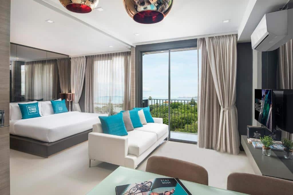 ห้องพักในโรงแรมทันสมัยพร้อมเตียงขนาดใหญ่ โซฟาสีขาว ทีวีจอแบน โต๊ะทำงาน และประตูกระจกที่เปิดออกไปสู่ระเบียงที่มองเห็นทิวทัศน์ของต้นไม้เขียวขจีและน้ำ เหมาะสำหรับการเข้าพักที่ผ่อนคลายที่ ที่พักพัทยา.