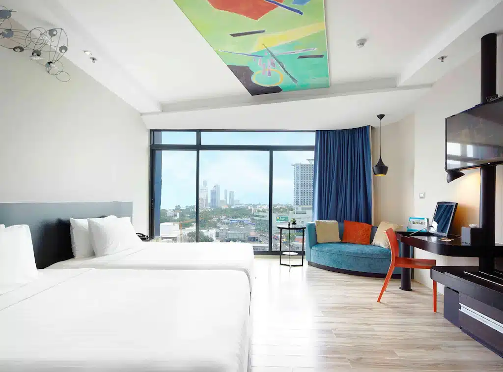 ห้องพักโรงแรมทันสมัยพร้อมเตียง 2 เตียง หน้าต่างบานใหญ่มองเห็นวิวเมือง โซฟาโค้งสีฟ้า ทีวีจอแบน โต๊ะพร้อมเก้าอี้สีแดง และศิลปะบนเพดานแบบนามธรรม เหมาะสำหรับผู้ที่มองหาที่พักพัทยาราคาถูกแต่มีสไตล์ในที่พักพัทยา.