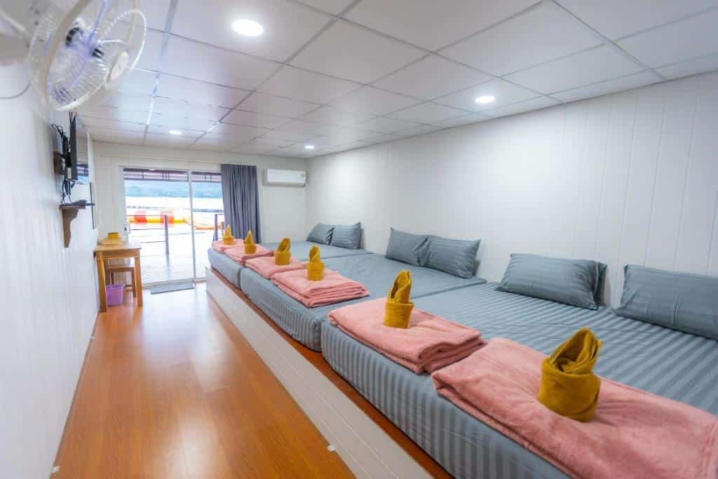 ห้องพักที่รีสอร์ทกาญจนบุรีมีเตียงสี่เตียงที่จัดเรียงอย่างเรียบร้อยพร้อมเครื่องนอนสีเทา ผ้าเช็ดตัวสีชมพู และผ้าเช็ดตัวสีเหลืองพับไว้ ติดกับทีวีติดผนังและประตูบานเลื่อนกระจกที่เปิดออกสู่ระเบียง—เหมาะสำหรับการพักผ่อนริมแม่น้ำของคุณในกาญจนบุรี.