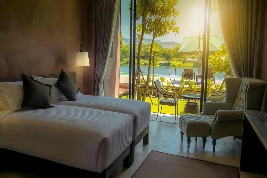 ห้องพัก ที่พักกาญจนบุรี โรงแรมสมัยใหม่พร้อมเตียงคู่สองเตียง, เก้าอี้นั่งเล่น, และหน้าต่างบานใหญ่ที่มองเห็นลานระเบียงพร้อมที่นั่งกลางแจ้งและพื้นที่สีเขียว—เหมาะสำหรับการพักผ่อนที่รีสอร์ทกาญจนบุรี พร้อมวิวสระว่ายน้ำหรือทะเลสาบและภูเขาเบื้องหลัง.