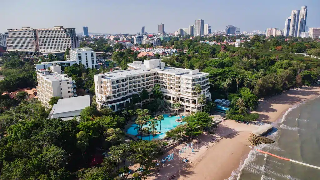 วิวทางอากาศของโรงแรมริมชายหาด หนึ่งในที่พักพัทยาราคาถูก ล้อมรอบด้วยต้นไม้ มีสระว่ายน้ำขนาดใหญ่ บริเวณชายหาด และมีเส้นขอบฟ้าของเมืองเป็นฉากหลัง.
