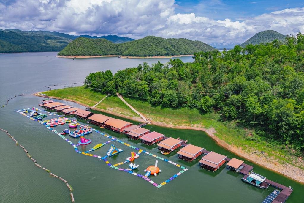 ภาพมุมสูงของบ้านลอยน้ำและเครื่องเล่นเป่าลมสีสันสดใสบนทะเลสาบที่ล้อมรอบด้วยเนินเขาสีเขียวและป่าไม้ใต้ท้องฟ้าที่มีเมฆบางส่วน เหมาะสำหรับการพักผ่อนที่ รีสอร์ทกาญจนบุรี หรือที่พักกาญจนบุรีริมน้ำ.