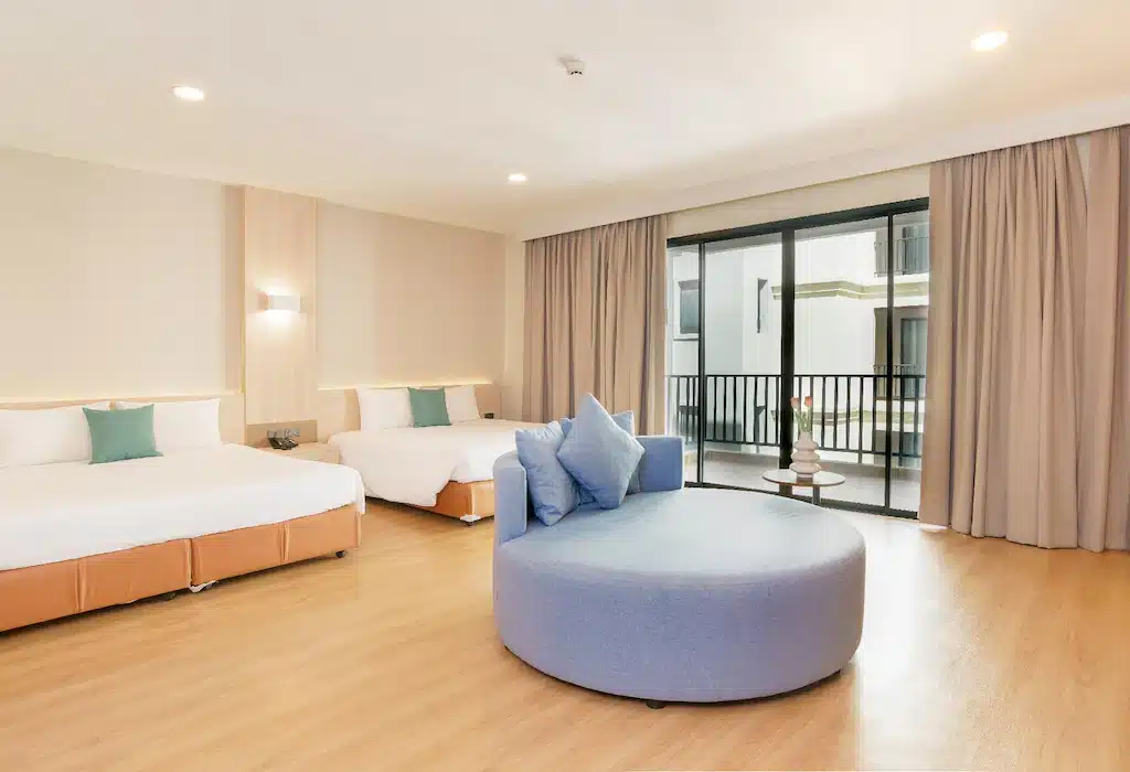 ห้องพักในโรงแรมกว้างขวางพร้อมเตียงคู่ 2 เตียง โซฟาทรงกลมสีฟ้า พื้นไม้ หน้าต่างบานใหญ่พร้อมผ้าม่าน และระเบียงเล็กๆ เหมาะสำหรับผู้ที่กำลังมองหาที่พักราคาถูกสำหรับการพักผ่อนอย่างผ่อนคลาย.