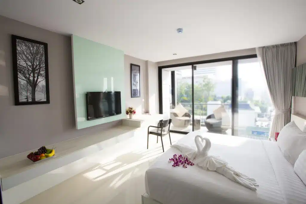 ห้องพักในโรงแรมทันสมัยพร้อมเตียงนอนที่จัดอย่างประณีต ผ้าขนหนูลายหงส์ กลีบดอกไม้ ทีวีจอแบน ชามผลไม้ และหน้าต่างบานใหญ่ที่เปิดสู่ระเบียงพร้อมวิวเมือง เหมาะสำหรับผู้ที่ต้องการพักในพัทยาเพื่อการพักผ่อนที่ผ่อนคลาย.