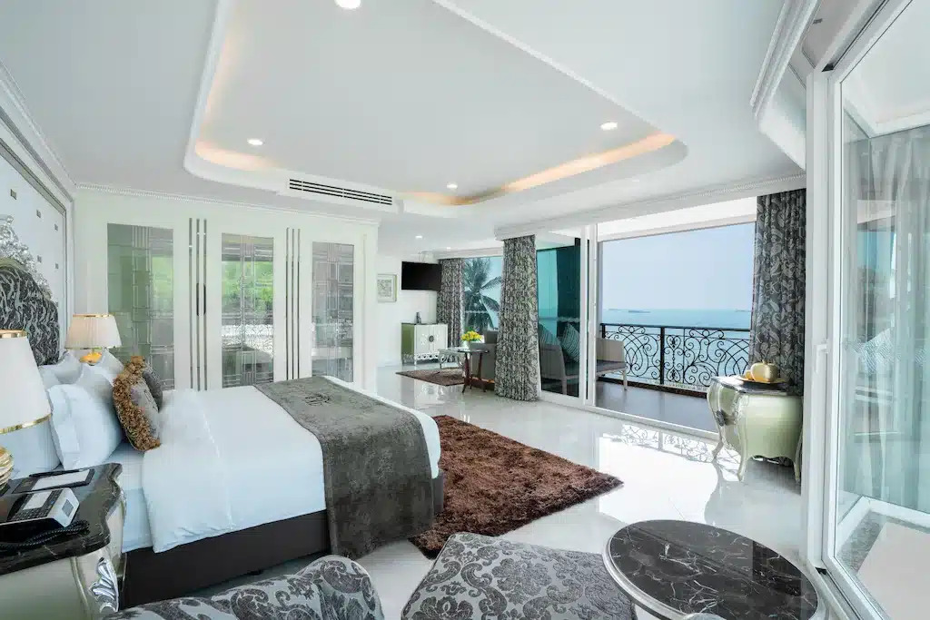 ห้องพักในโรงแรมทันสมัย พร้อมเตียงขนาดใหญ่ พรมหรูหรา พื้นที่นั่งเล่น ทีวีจอแบน และระเบียงที่มองเห็นวิวทะเล เหมาะสำหรับนักเดินทางที่กำลังมองหาที่พักพัทยาราคาถูกพร้อมการตกแต่งที่หรูหราด้วยสีขาวและสีเงิน.