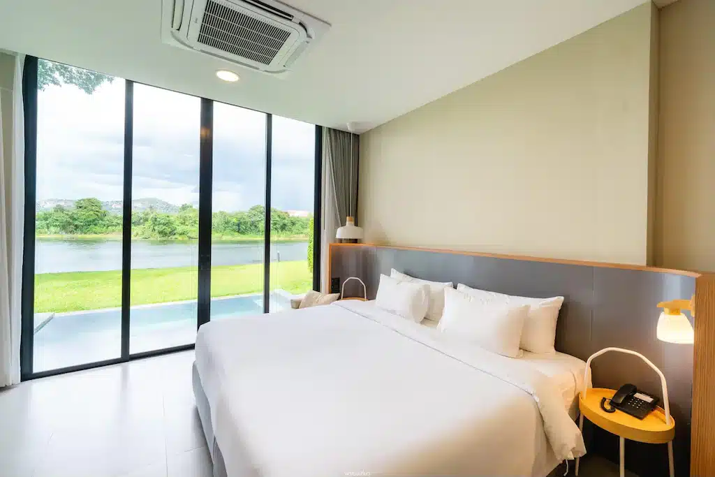 ห้องพักโรงแรมสมัยใหม่ในรีสอร์ท ที่พักกาญจนบุรี กาญจนบุรี พร้อมเตียงขนาดใหญ่ โต๊ะข้างเตียงสองตัวพร้อมโคมไฟ หน้าต่างสูงจากพื้นจรดเพดาน และวิวที่สวยงามของแม่น้ำและต้นไม้เขียวขจีภายนอก.
