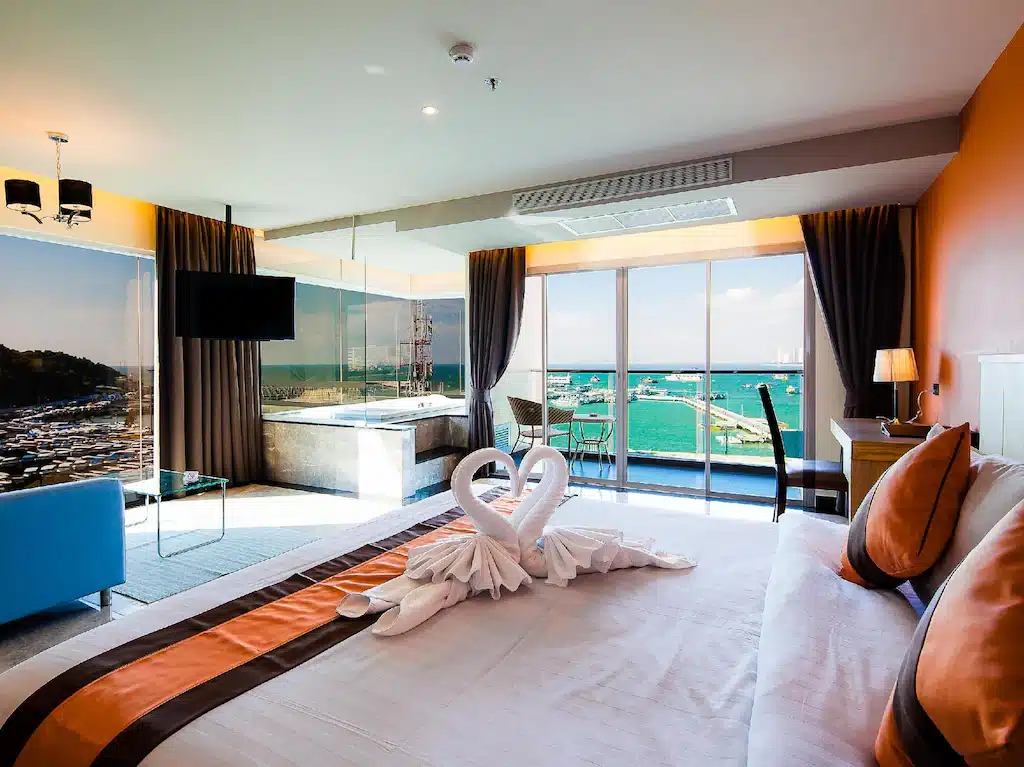 ห้องพักในโรงแรมทันสมัยพร้อมหน้าต่างบานใหญ่ที่มองเห็นท่าจอดเรือ เตียงพร้อมผ้าเช็ดตัวลายหงส์ อ่างอาบน้ำข้างหน้าต่าง โต๊ะทำงาน และทีวีจอแบน เหมาะสำหรับผู้ที่ต้องการที่พักในพัทยาที่มีสไตล์และสะดวกสบาย.
