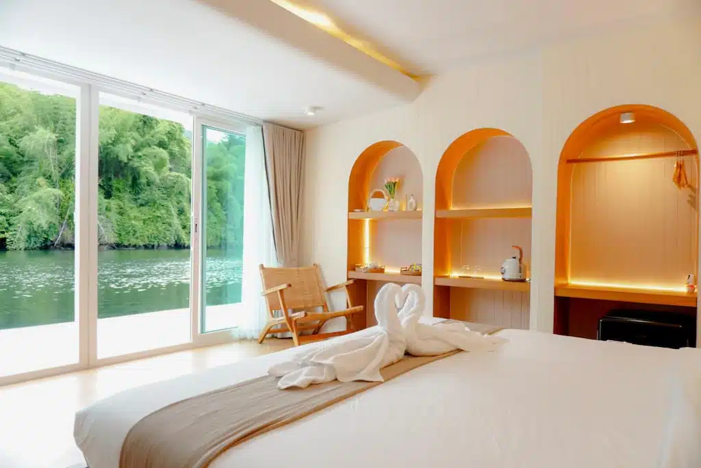 ห้องพักโรงแรมสมัยใหม่พร้อมเตียงขนาดใหญ่ ผ้าเช็ดตัวรูปหงส์ ชั้นวางของในตัว เก้าอี้ และหน้าต่างบานใหญ่จากพื้นจรดเพดานที่มองเห็นแม่น้ำและธรรมชาติสีเขียว—เหมาะสำหรับการเข้าพักของคุณที่ ที่พักกาญจนบุรีริมน้ำ 