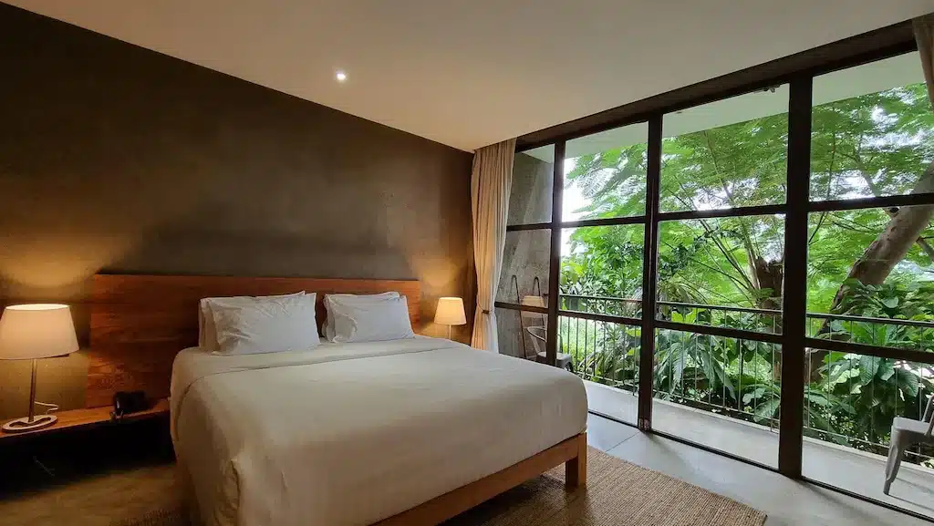 ห้องนอนสไตล์โมเดิร์นในรีสอร์ท ที่พักกาญจนบุรี กาญจนบุรี พร้อมเตียงคู่ หัวเตียงไม้ โคมไฟข้างเตียง และหน้าต่างบานใหญ่จากพื้นจรดเพดานที่มองเห็นวิวทิวทัศน์สีเขียวขจี.