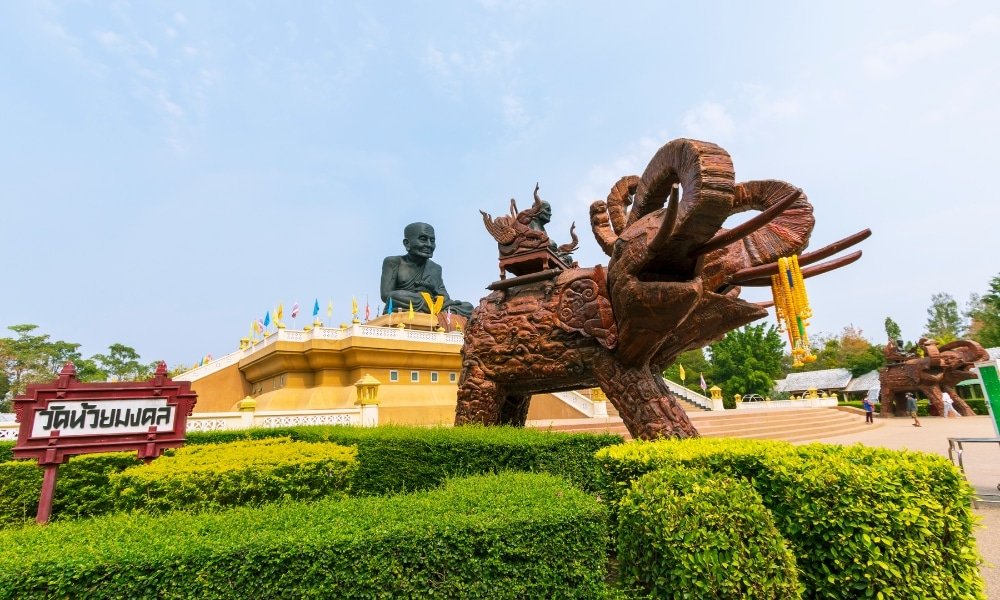 รูปปั้นพระภิกษุสัมฤทธิ์ขนาดใหญ่ตั้งอยู่บนแท่นสีทองด้านหลังรูปปั้นช้างอันหรูหราที่วัดแห่งหนึ่งใน ท่องเที่ยวหัวหิน ประเทศไทย ซึ่งเป็นหนึ่งในไฮไลท์ของท่องเที่ยวหัวหิน โดยมีป้ายและพุ่มไม้ที่ตกแต่งอย่างสวยงามอยู่เบื้องหน้า.