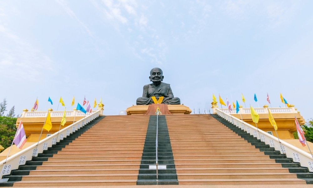 บันไดหินกว้างทอดไปสู่รูปปั้นชายร่างใหญ่นั่งขนาบข้างด้วยราวสีเหลือง ท่องเที่ยวหัวหิน และธงหลากสีสันใต้ท้องฟ้าแจ่มใส สถานที่ที่นักท่องเที่ยวไม่ควรพลาดในการสำรวจท่องเที่ยวหัวหิน.