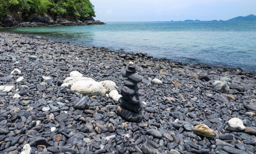 กองหินสีดำเรียบวางซ้อนกันอยู่บนชายหาดหินริมทะเล เป็นภาพที่เงียบสงบ ณ สถานที่ท่องเที่ยวธรรมชาติ ท่องเที่ยวสตูล หัวหิน โดยมีต้นไม้สีเขียวและเกาะไกลๆ เป็นฉากหลัง ภายใต้ท้องฟ้าที่มีเมฆบางส่วน.