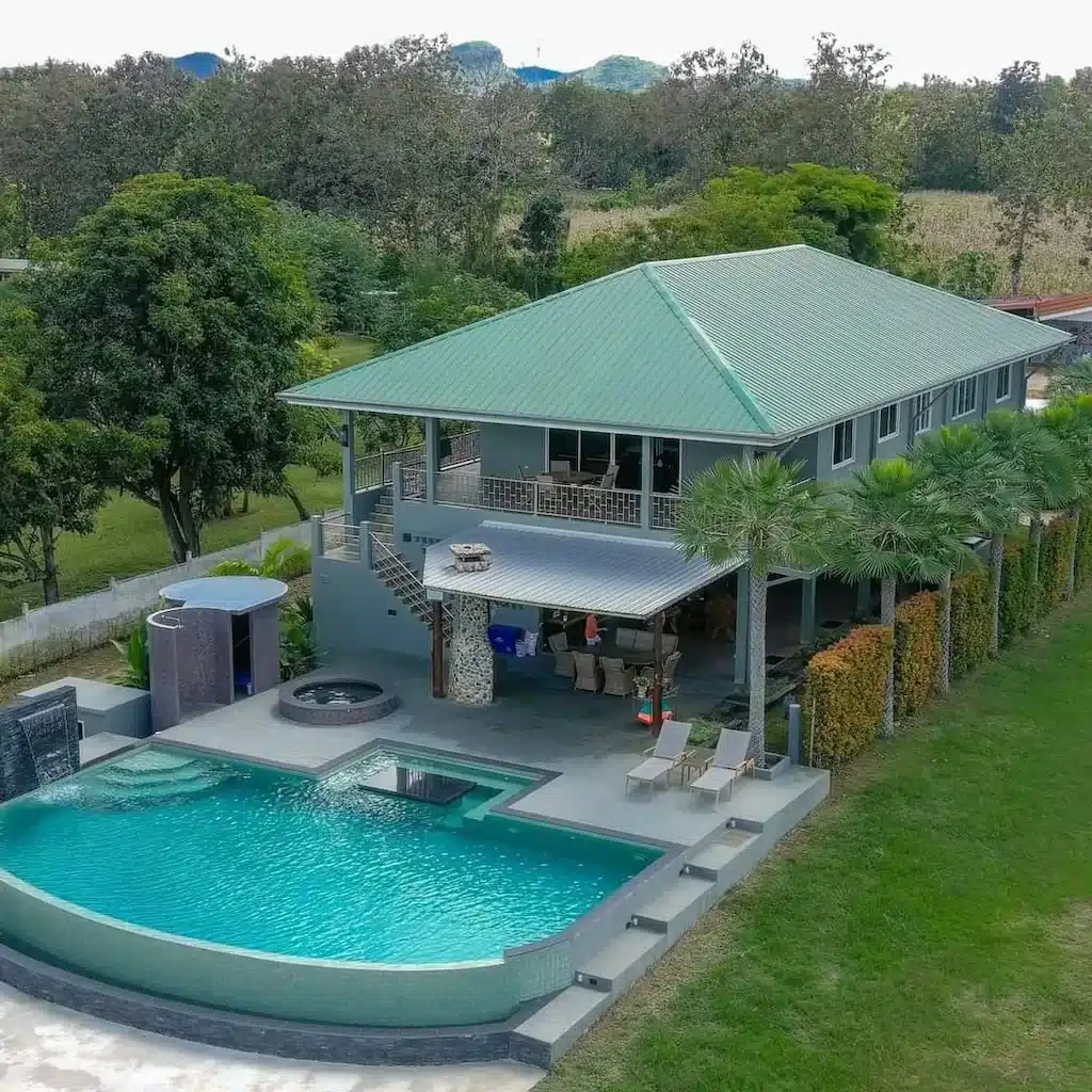 บ้านสมัยใหม่ที่มีหลังคาสีเขียวและบันไดภายนอก พร้อมสระว่ายน้ำ, อ่างน้ำร้อน, เก้าอี้อาบแดดริมสระ และ ที่พักกาญจนบุรี ล้อมรอบด้วยต้นไม้—เหมาะสำหรับการพักผ่อนที่ที่พักกาญจนบุรีริมน้ำหรือรีสอร์ทกาญจนบุรี.