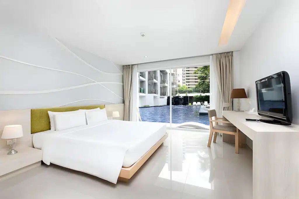 ห้องพักโรงแรมทันสมัยในพัทยาราคาถูก มีเตียงขนาดใหญ่ โต๊ะทำงาน ทีวีจอแบน และประตูกระจกบานเลื่อนพร้อมวิวสระว่ายน้ำ การตกแต่งเรียบง่ายด้วยผนังสีขาวและเฟอร์นิเจอร์สีอ่อน เหมาะสำหรับการเข้าพักของคุณ.