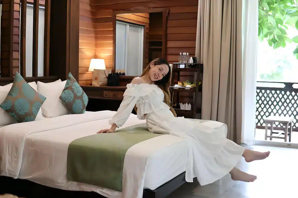 ผู้หญิงในชุดสีขาว นั่งอยู่บนขอบเตียงที่จัดไว้อย่างเรียบร้อย ในห้อง รีสอร์ทกาญจนบุรี ที่อบอุ่นและสว่างไสว ผนังไม้ และตกแต่งอย่างทันสมัย.