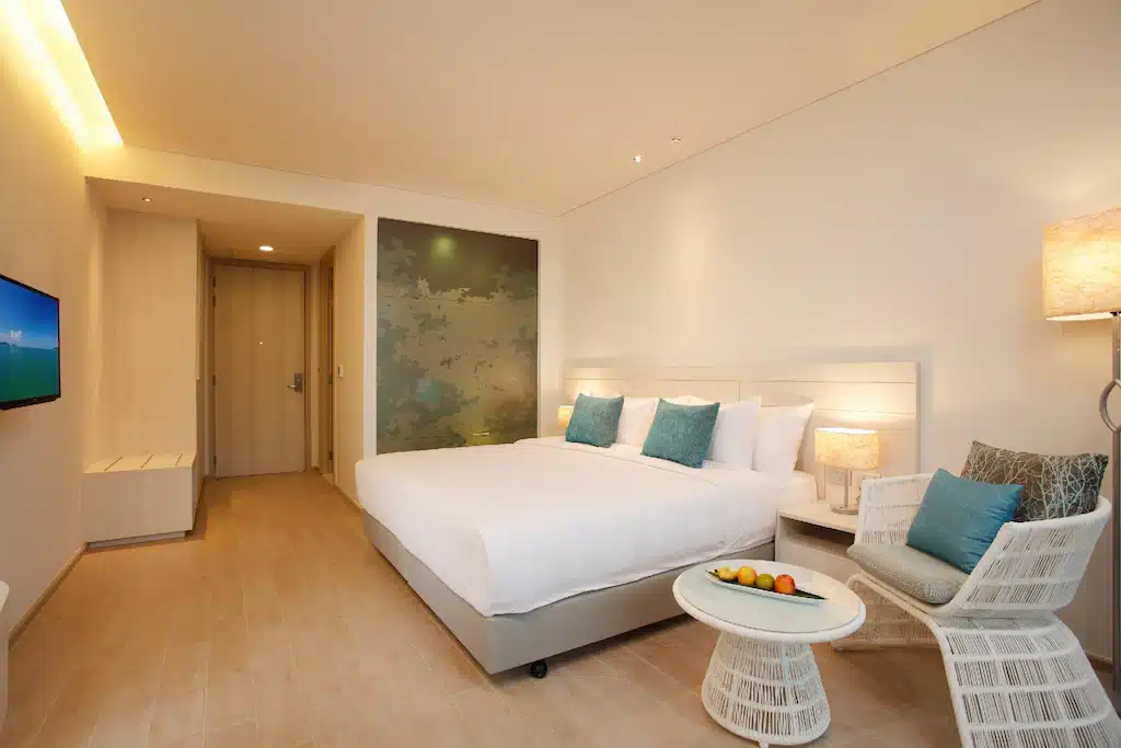 ห้องพักในโรงแรมทันสมัยพร้อมเตียงขนาดใหญ่ หมอนสีฟ้า โต๊ะข้างพร้อมผลไม้ เก้าอี้สีขาว และทีวีติดผนัง เพลิดเพลินไปกับการตกแต่งแบบเรียบง่ายและแสงไฟอันอบอุ่นที่โรงแรมในพัทยาแห่งนี้ซึ่งเหมาะสำหรับการเข้าพักของคุณ.