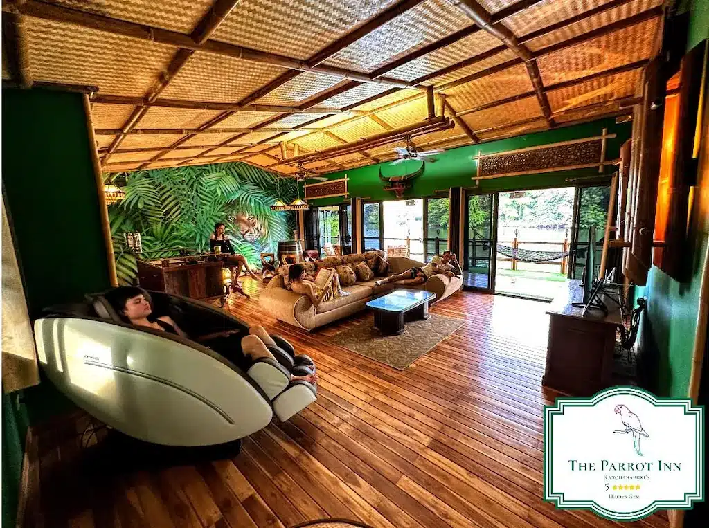 เลานจ์กว้างขวางในธีมเขตร้อนที่ The Parrot Inn มีพื้นไม้ เพดานหวาย ผนังสีเขียว หน้าต่างบานใหญ่ และเก้าอี้นวด—เหมาะอย่างยิ่งสำหรับการผ่อนคลายที่ ที่พักกาญจนบุรีริมน้ำ แห่งนี้.
