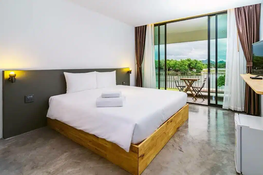 ห้องพักโรงแรมสไตล์มินิมอลพร้อม ที่พักกาญจนบุรี เตียงคู่, โคมไฟข้างเตียง, โต๊ะทำงานขนาดเล็ก, โทรทัศน์จอแบน, และระเบียงที่มองเห็นวิวสีเขียวและน้ำ—เหมาะสำหรับผู้ที่ต้องการที่พักริมน้ำกาญจนบุรีหรือประสบการณ์รีสอร์ทกาญจนบุรีที่ผ่อนคลาย.