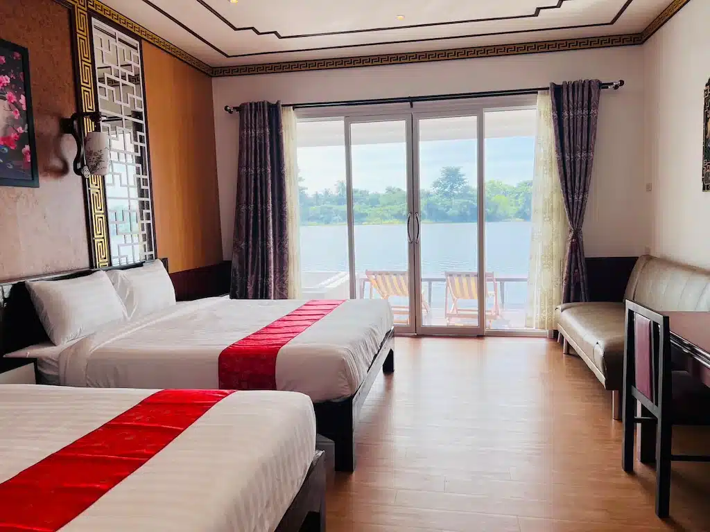 ห้องพักโรงแรม ที่พักกาญจนบุรี พร้อมเตียงคู่สองเตียง พรมปูโต๊ะสีแดง โซฟา โต๊ะทำงาน และประตูกระจกที่นำไปสู่ระเบียงพร้อมวิวแม่น้ำและเก้าอี้อาบแดด—ที่พักริมแม่น้ำกาญจนบุรีที่เหมาะสำหรับการพักผ่อนของคุณ.