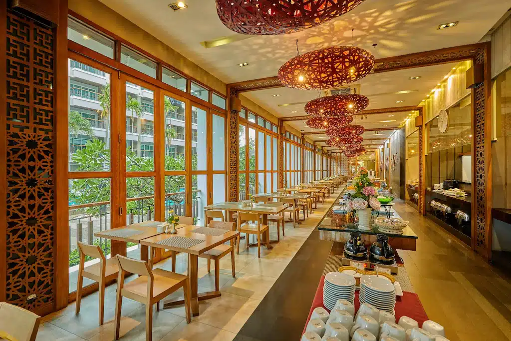 พื้นที่รับประทานอาหารในร้านอาหารที่ยาวและมีแสงสว่างเพียงพอ พร้อมโต๊ะและเก้าอี้ไม้ หน้าต่างบานใหญ่ ไฟเพดานสีแดงอันวิจิตรบรรจง และการจัดบุฟเฟ่ต์ไว้ตามผนังด้านหนึ่ง เหมาะสำหรับแขกที่กำลังมองหาที่พักพัทยาราคาถูก และประสบการณ์การรับประทานอาหารอันอบอุ่น.