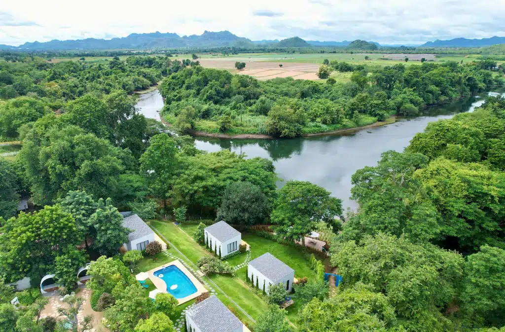 ภาพมุมสูงของกระท่อมสีขาวขนาดเล็กและสระว่ายน้ำล้อมรอบด้วยต้นไม้ติด ที่พักกาญจนบุรี แม่น้ำ มอบที่พักริมน้ำแคนจันบุรีที่เงียบสงบ พร้อมทุ่งนาและภูเขาเป็นฉากหลัง.